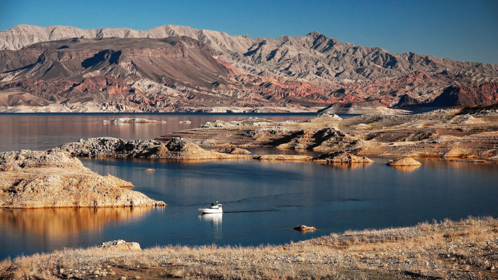A small white motorboat cruises across the calm, dark blue water of Lake Mead, surrounded by rugged desert terrain. In the foreground, rocky, sun-bleached peninsulas and small islands dot the water's edge. The background is dominated by massive, colorful mountain ranges with visible layers of red, tan, and gray rock under a clear, bright sky.