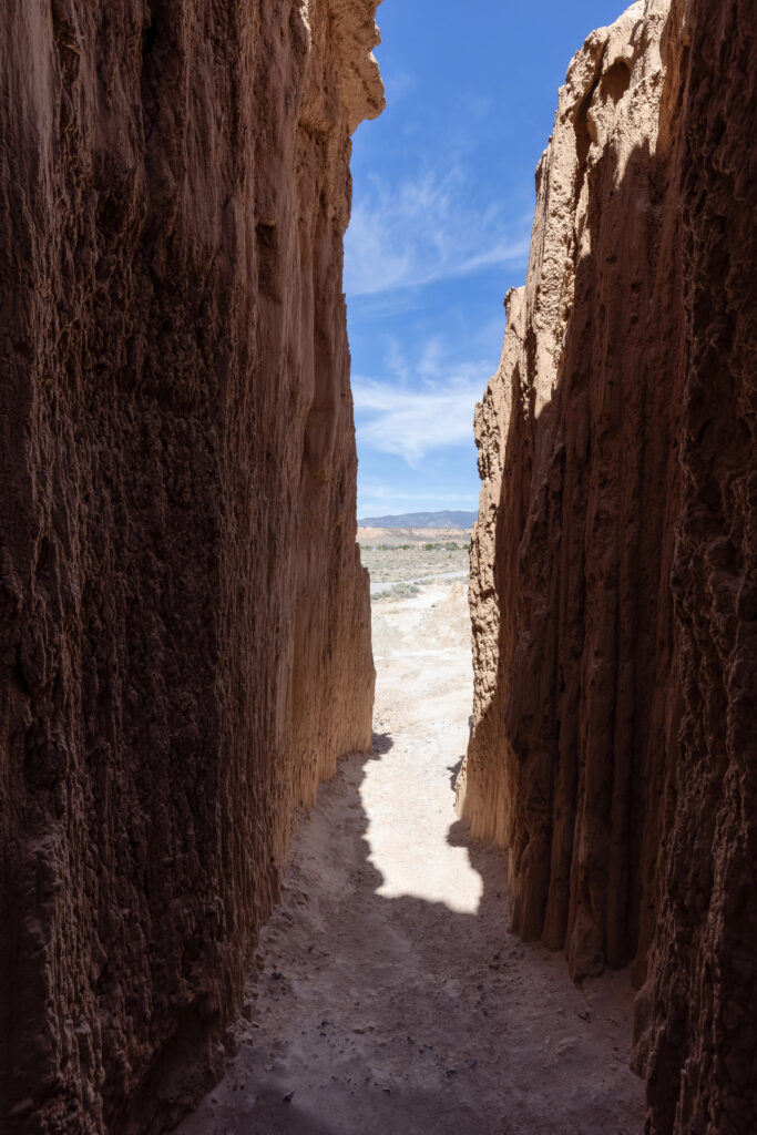 A narrow, vertical view from inside a deep slot canyon made of tan, eroded bentonite clay. The sheer, textured walls frame a bright opening that reveals a sunlit desert floor and distant blue mountains under a light sky. The foreground is in deep shadow, emphasizing the tight, cathedral-like passage of the "moon caves."