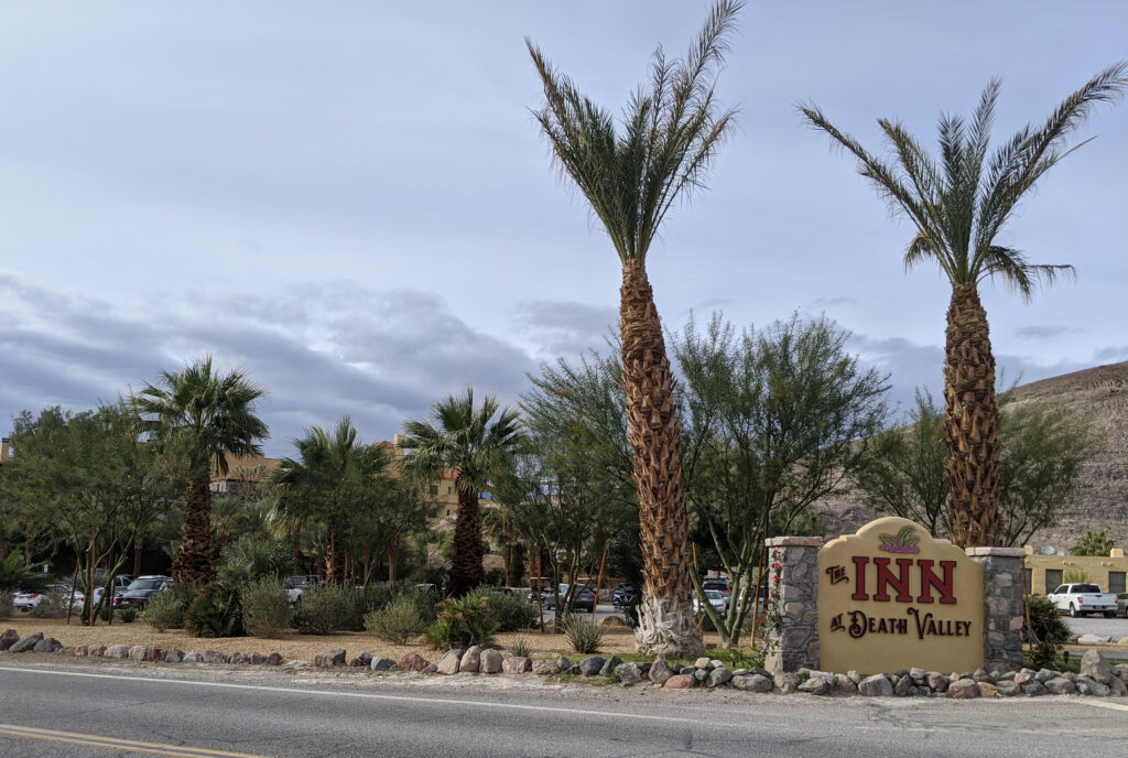 An exterior view of The Inn at Death Valley, a historic mission-style resort built into a rocky hillside. The building features warm-toned stone walls, red-tiled roofs, and elegant arches. Lush greenery, including towering date palms and colorful bougainvillea, surrounds the property, creating a vibrant oasis against the stark, arid desert mountains in the background.