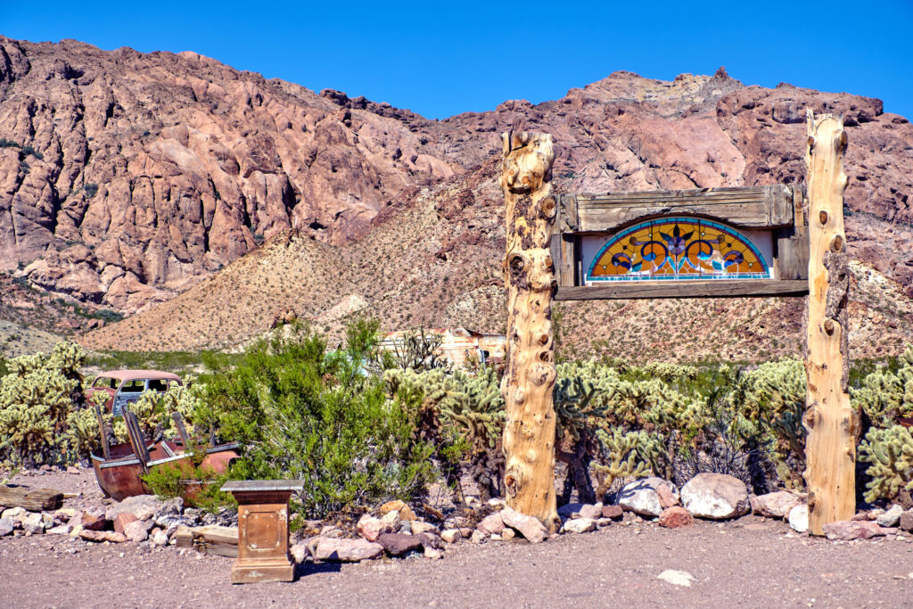 A rustic scene at Nelson Ghost Town featuring a cluster of weathered wooden buildings and vintage relics under a bright desert sky. In the foreground, several rusted antique cars from the early 20th century are parked on the dusty ground. The backdrop includes rugged, rocky desert hills and a collection of eclectic items like old gasoline pumps, weathered signs, and the remnants of a wooden mining structure.