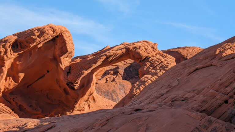 A curved red sandstone arch rises from layered rock formations in Valley of Fire State Park, glowing against a bright blue desert sky.