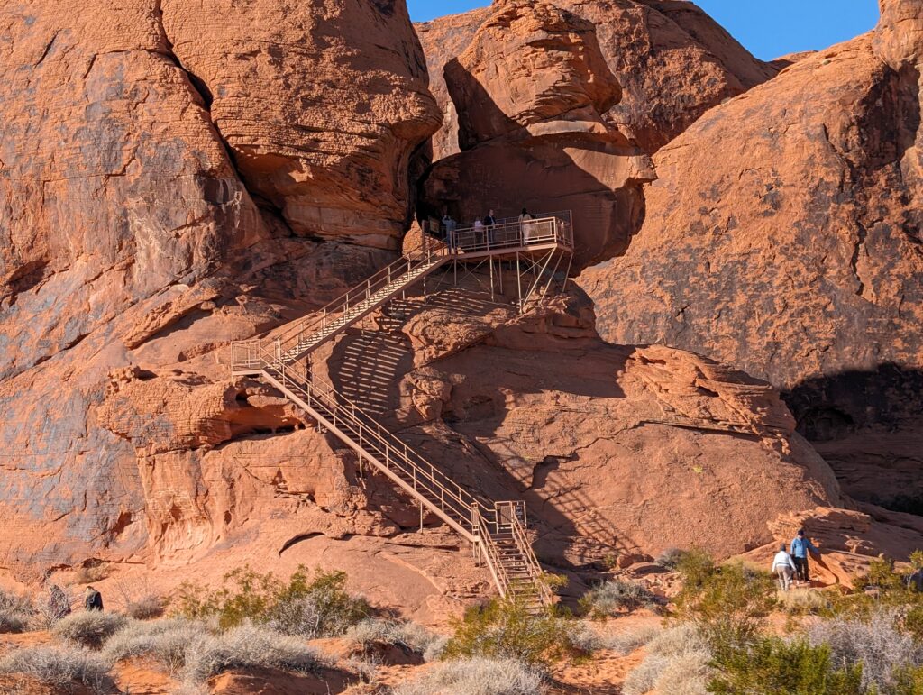 A towering, vibrant orange Aztec Sandstone formation under a clear blue sky at Atlatl Rock in Valley of Fire State Park. A long, tan metal staircase with handrails zigzags up the side of the steep, rugged rock face, leading to a viewing platform where several people are standing.