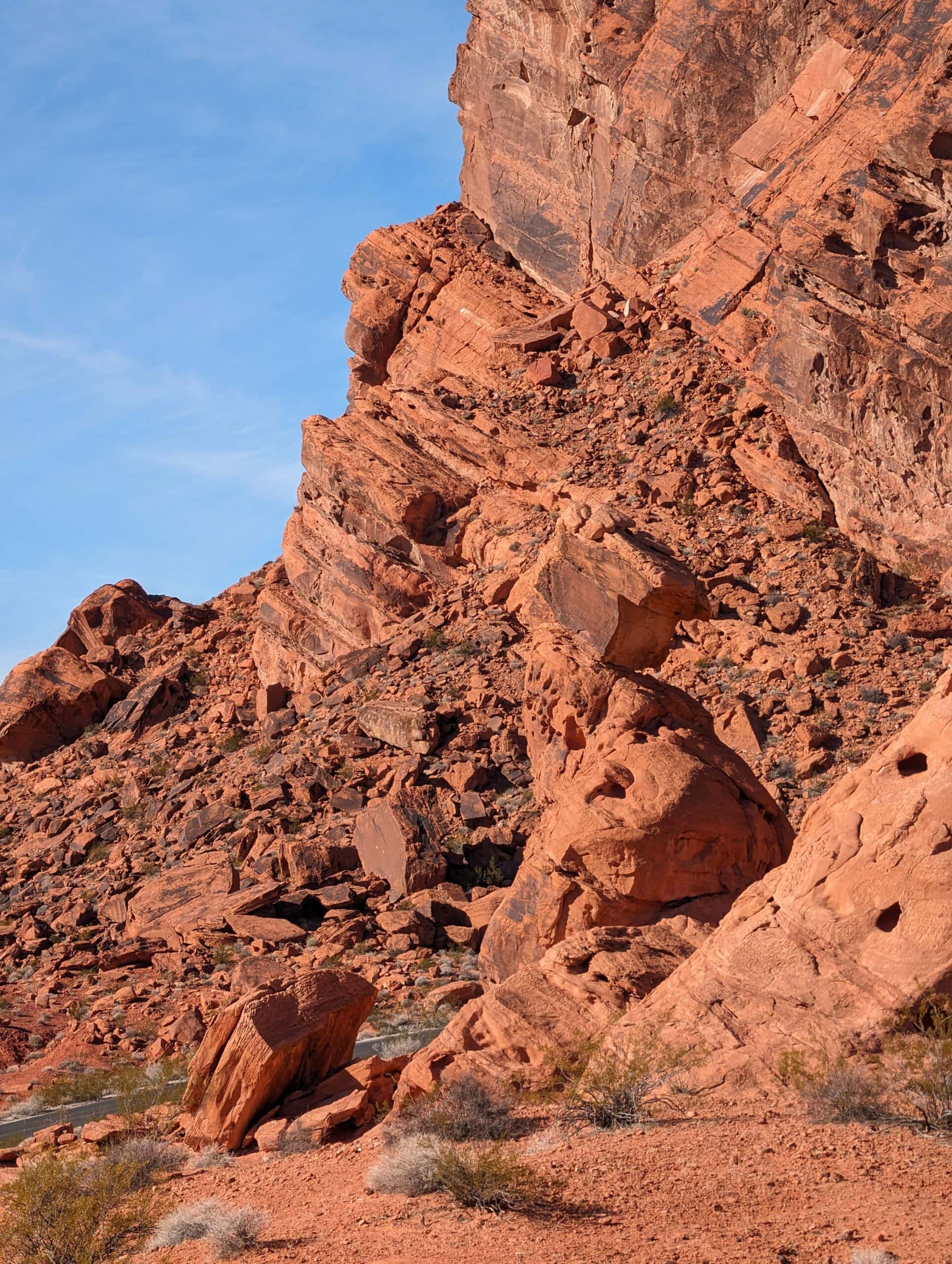 A vertical shot of the "Balancing Rock" formation in Valley of Fire State Park. The centerpiece is a large, rectangular red sandstone boulder precariously perched atop a narrow, tapering rock pedestal. The surrounding landscape is a steep hillside of jagged, tumbled orange-red rocks and boulders under a pale blue sky. In the lower left corner, a small section of a paved gray road is visible, providing a sense of scale to the massive rock formations.