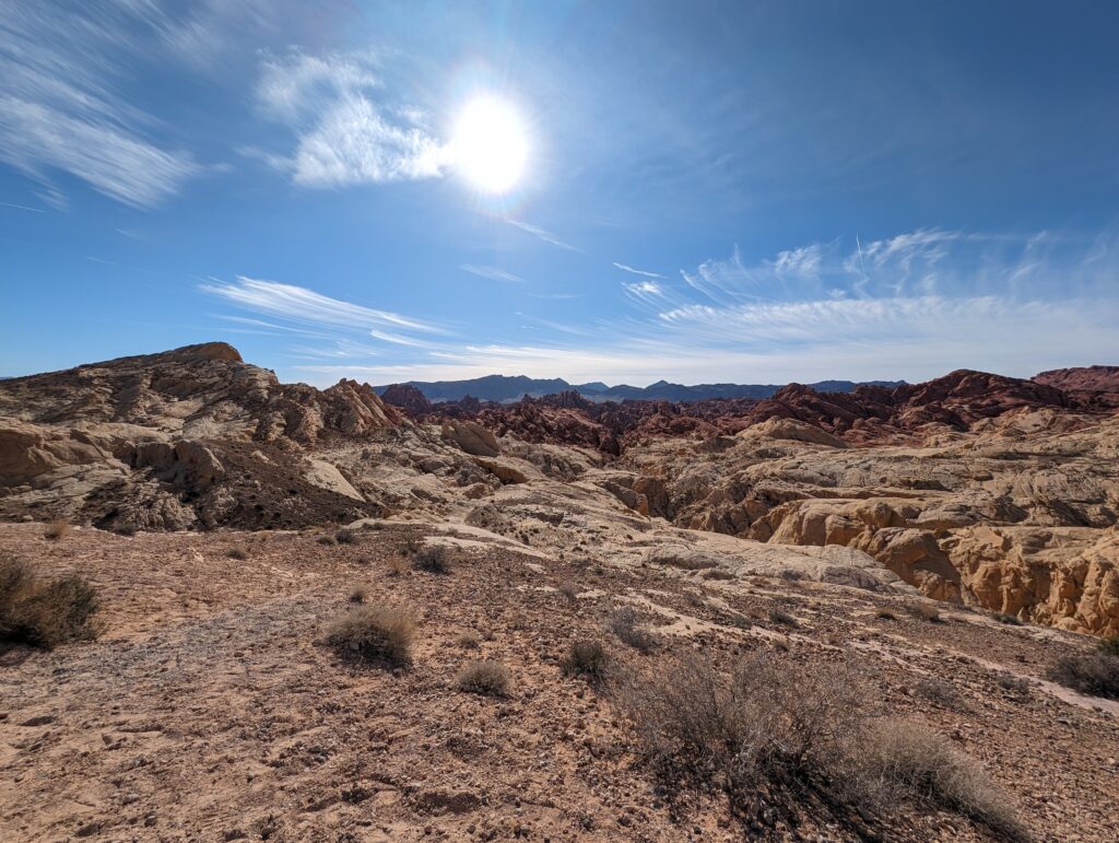 Wide desert landscape at the Fire Canyon and Silica Dome viewpoint in Valley of Fire State Park, with pale sandstone hills, red rock ridges, and distant mountains under a bright blue sky.