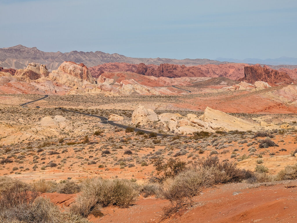 Wide view of distant road winding through layered red rock hills and desert landscape at Valley of Fire State Park under a hazy blue sky.