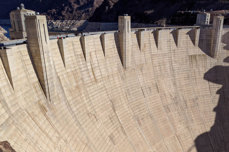 The curved concrete face of Hoover Dam fills the view with visitors walking along the top. The massive scale is a highlight of a Hoover Dam day trip from Las Vegas.