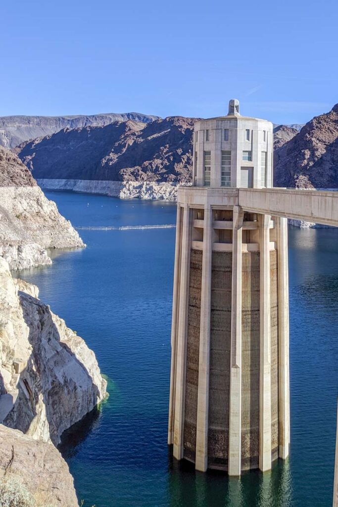 A tall concrete intake tower rises from the deep blue water of Lake Mead beside rocky desert cliffs. The visible pale shoreline shows how far the water level has fallen.