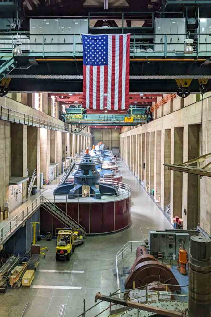 Rows of large hydroelectric generators extend through the Hoover Dam power plant beneath a hanging American flag. The cavernous turbine hall is one of the main indoor stops on the guided tours.