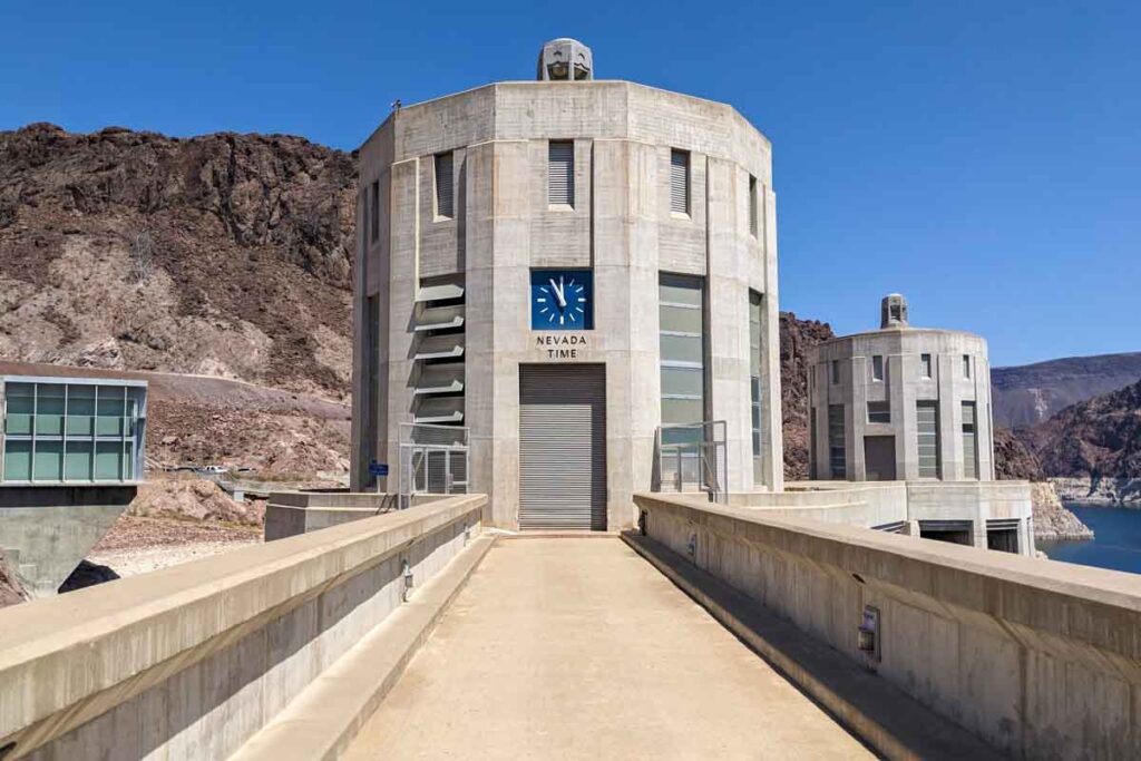 Two concrete intake towers stand beside Lake Mead under a clear blue desert sky. A clock on the nearest tower reads "Nevada Time" and marks the state line experience during a Hoover Dam day trip from Las Vegas.