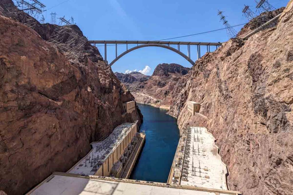 The Colorado River runs between steep Black Canyon walls below the Mike O Callaghan Pat Tillman Memorial Bridge. Power plant buildings line the river at the base of Hoover Dam.