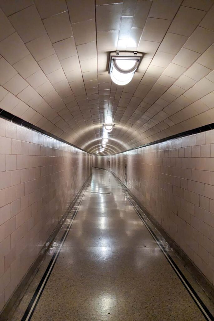 A long curved tunnel with tiled walls and bright ceiling lights leads through the interior of Hoover Dam. The polished walkway gives visitors a sense of the hidden spaces inside the structure.